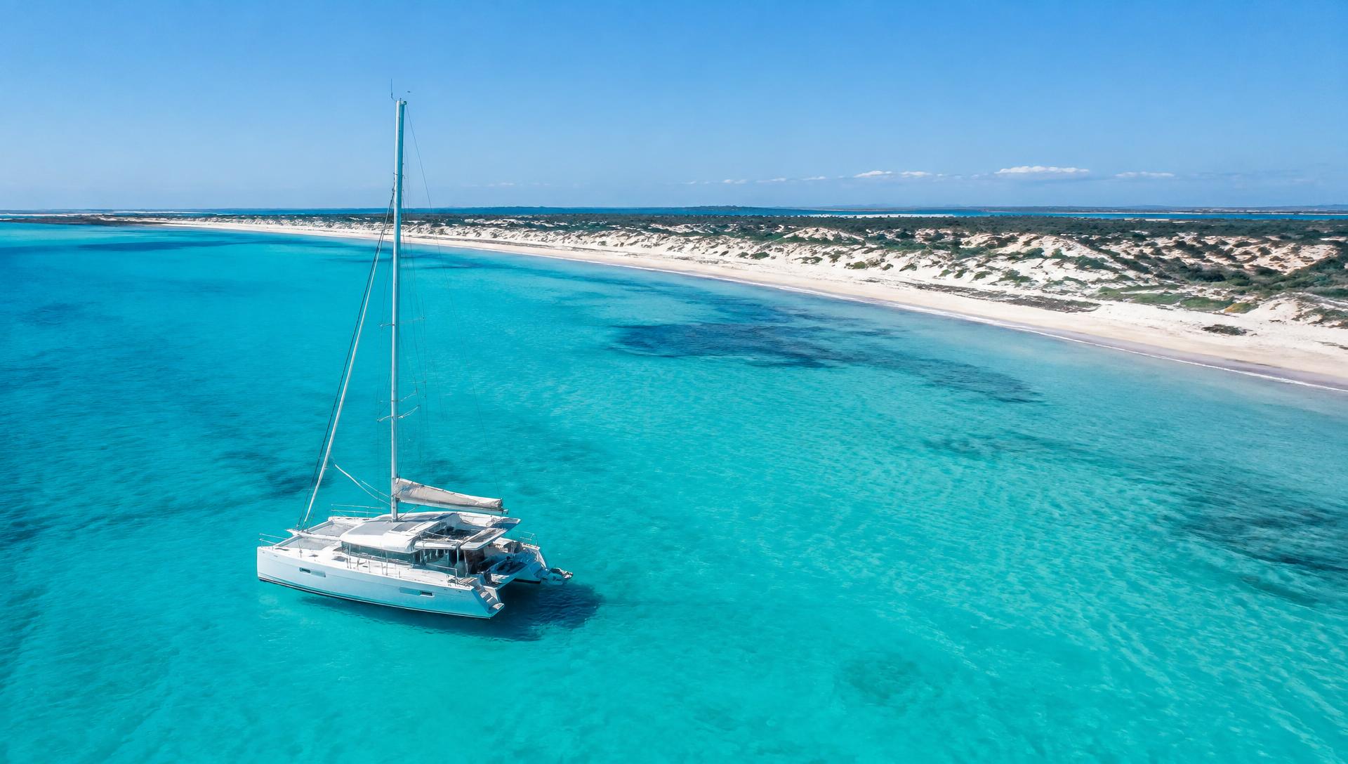 Sailing catamaran anchored over pale turquoise shallows off a long white sand beach in Formentera
